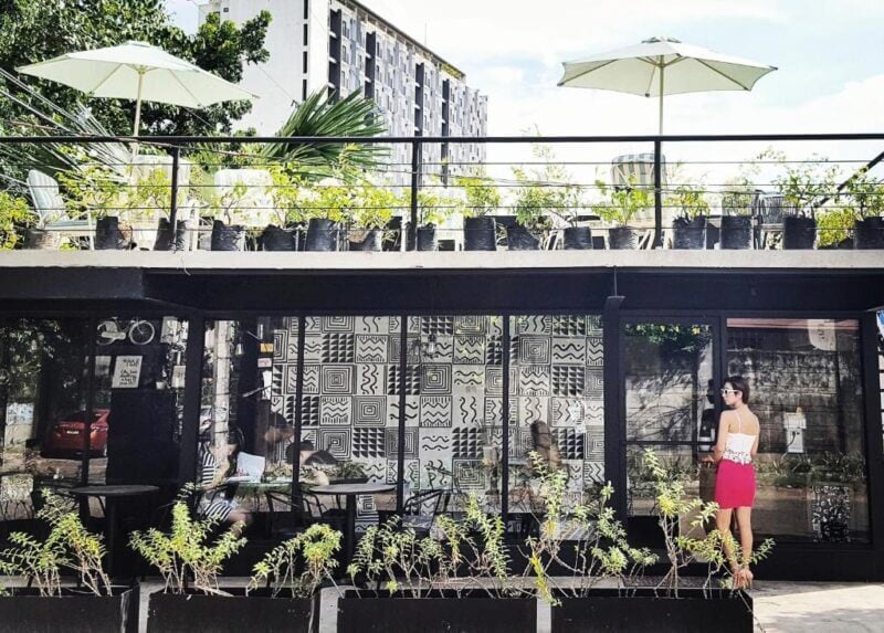 A woman in a white top and red skirt stands outside Murals Hostel Cebu, a modern café-like hostel with patterned walls, potted plants, and outdoor seating under umbrellas.