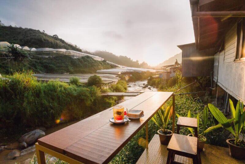 A wooden table with a drink and snack overlooks a small river and lush greenery at sunrise in Cameron Highlands, next to a building and potted plants—capturing the tranquil spirit of Heahtitude.