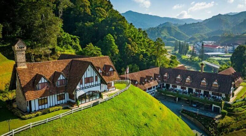 Aerial view of Tudor-style LakeHouse buildings with red roofs on a green hillside in Cameron Highlands, surrounded by trees and mountains under a partly cloudy sky.