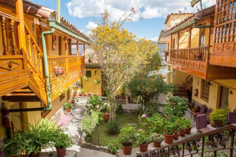 A sunny courtyard at Amaru Colonial Cusco features potted plants, trees, and flowers, all surrounded by charming yellow buildings with wooden balconies and railings.