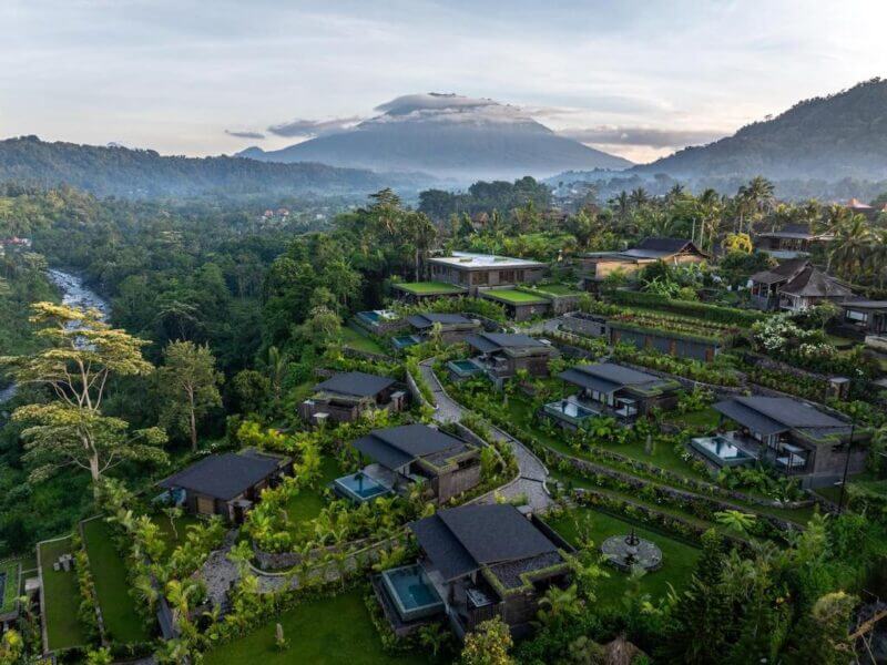 Aerial view of Samanvaya Resort in Sidemen, showcasing modern villas, pools, and gardens nestled on a lush hillside, surrounded by tropical forest and backed by a majestic mountain under a partly cloudy sky.