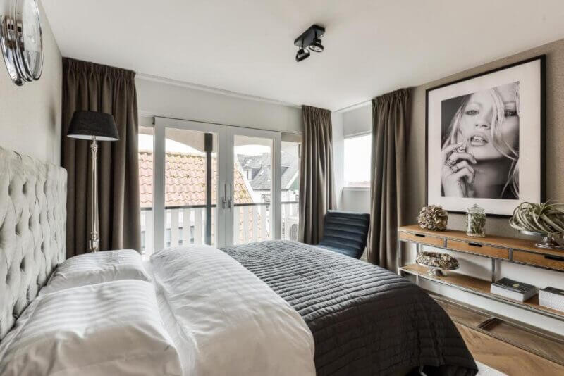 Modern bedroom with a double bed, gray quilt, large window doors, a black chair, wall art above a shelf, and neutral-toned curtains—perfect for a boutique hotel escape near The Old Bridge in Volendam.