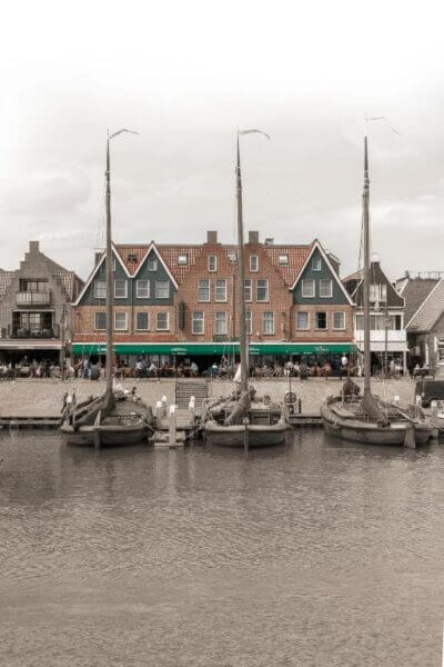 Three docked sailboats rest before traditional gabled buildings along the waterfront, with people strolling beside the charming Old Dutch and Volendam hotel in the background.