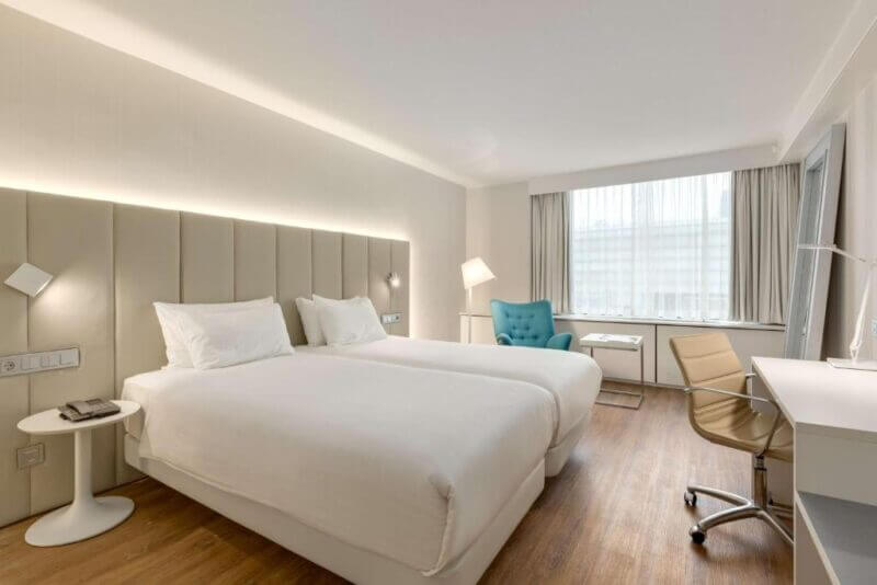 Modern Utrecht hotel room with two single beds, a white desk and chair, armchair by the window, wooden flooring, and neutral decor—perfect comfort in the heart of NH Utrecht.