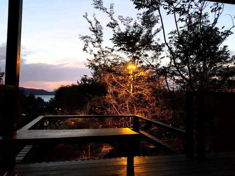 A wooden deck at Annie's Koh Yao Noi accommodation overlooks trees and a distant body of water at sunset, with a streetlamp softly illuminating the lush foliage.