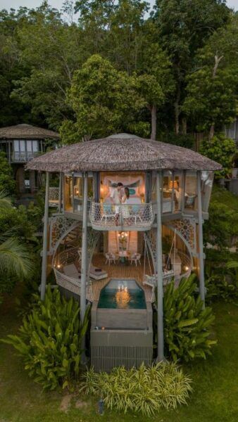 A couple stands on the balcony of a two-story TreeHouse Koh Yao Noi villa with a thatched roof, private pool, and surrounded by lush greenery.