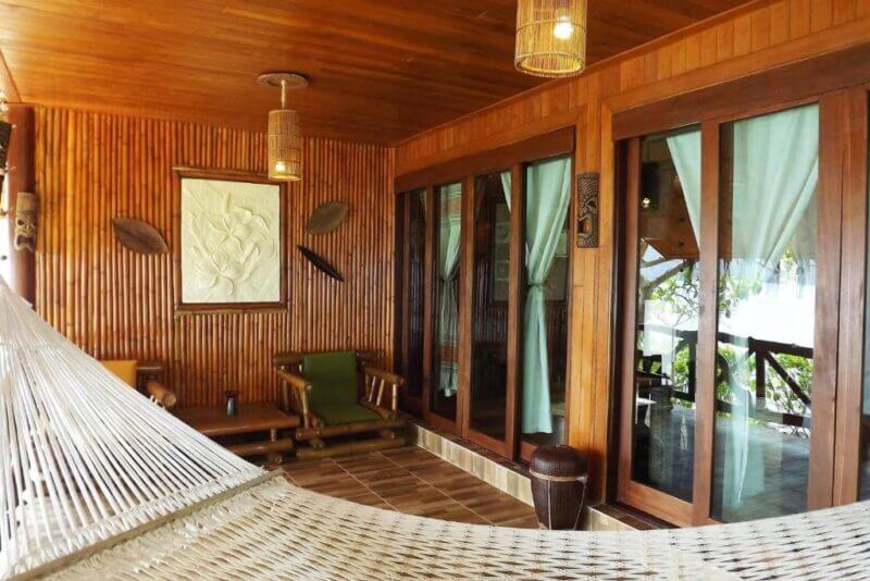 A wooden patio at Viking Resort Phi Phi features bamboo walls, glass sliding doors with curtains, a hammock, a cozy green-cushioned seating area, and ceiling lights.