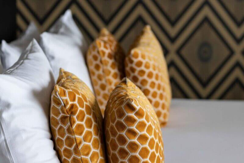 Four orange hexagonal patterned pillows and two white pillows are arranged on a bed at The James Rotterdam, with a geometric patterned wall in the background.