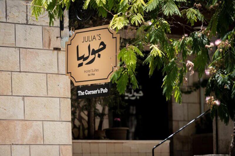 A sign reads Julia's Hotel Suites in English and Arabic in Amman, with The Corners Pub below, mounted on a stone wall by leafy branches.