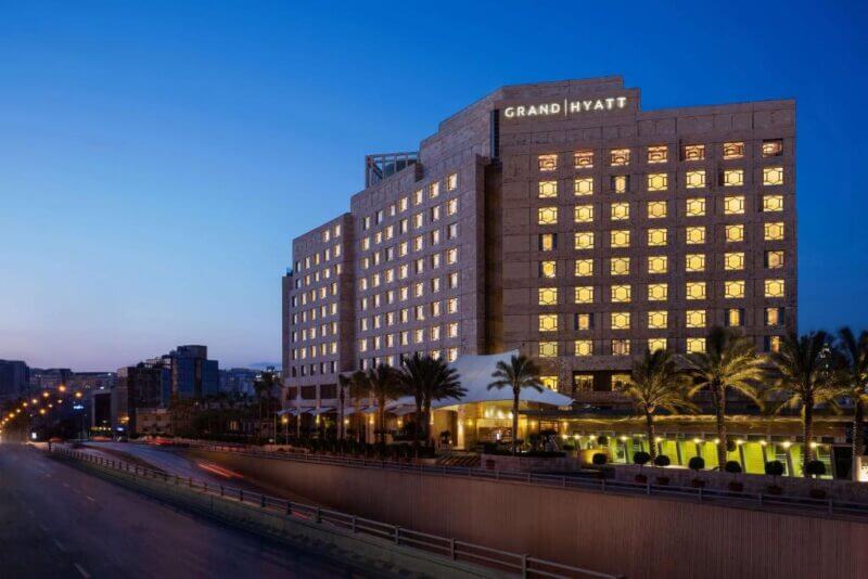 Exterior view of the Grand Hyatt Amman at dusk, with illuminated windows and palm trees lining the elegant entrance.