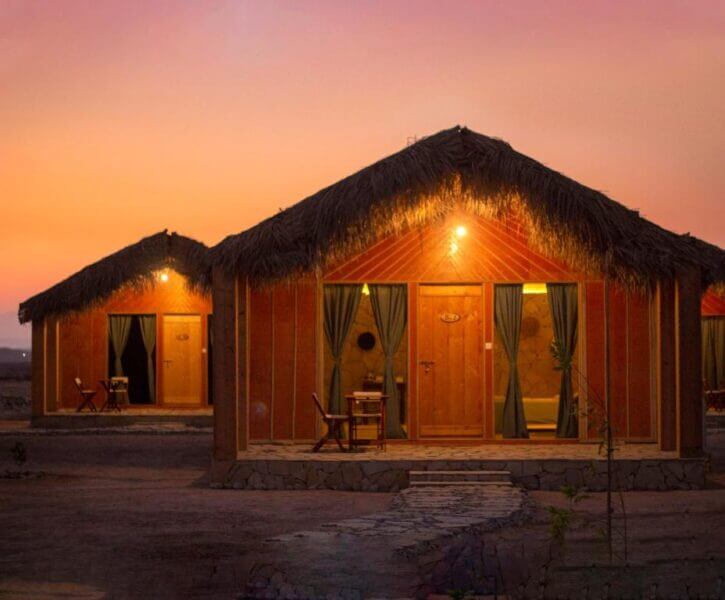 Two wooden huts with thatched roofs are illuminated at sunset at Sama Sur Resort, each with green curtains and outdoor seating, set on a stone-lined path in a dry landscape.