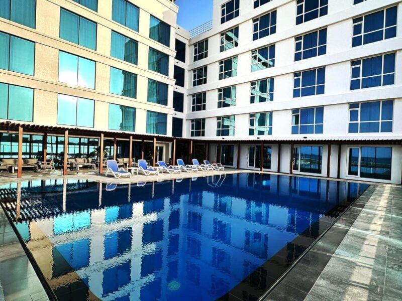Outdoor pool with blue water surrounded by lounge chairs, next to a grand hotel building featuring multiple stories and large windows.