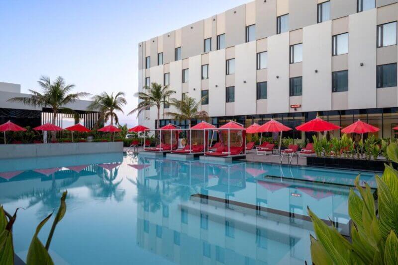 Hotel outdoor pool area at Avani Muscat with red lounge chairs and umbrellas, palm trees, and a modern three-story building in the background—perfect for relaxing at this stylish Oman hotel.