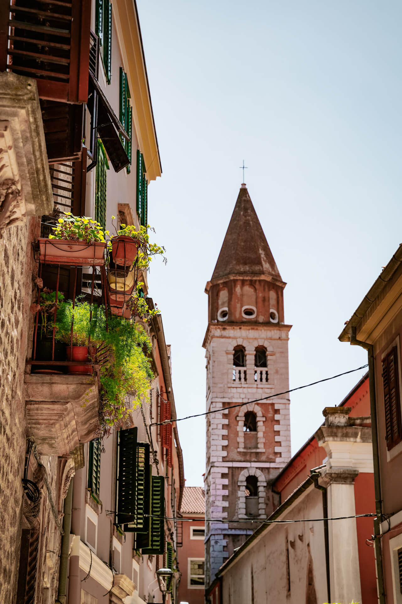 A stone bell tower with arched windows rises above the narrow streets of Zadar, Croatia, where buildings with green-shuttered windows bask in the sunny day.