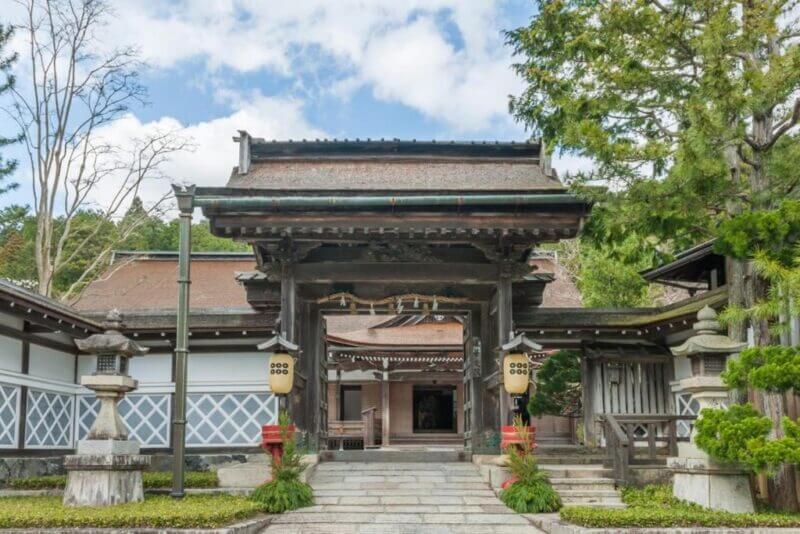 Traditional Japanese shrine entrance with a wooden gate, stone lanterns, and decorative greenery at Rengejoin Temple in Koyasan, set against trees and a partly cloudy sky.
