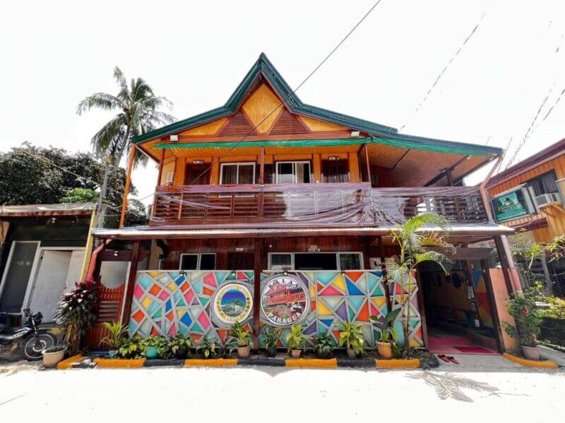 A two-story wooden building in El Nido with a geometric mural and plants in front, featuring a balcony and triangular roof, a palm tree on the left side, and inspired by Balay Paragua’s distinctive local charm.