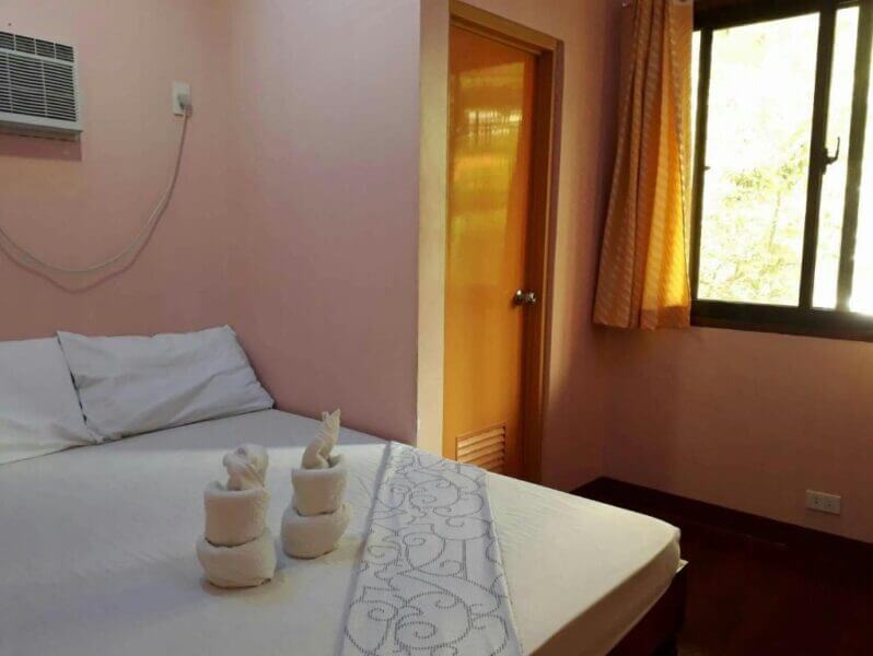 A neatly made bed with white linens and towel sculptures sits next to a window with yellow curtains and a closed door in a small room with peach-colored walls at David's Guest House in El Nido.
