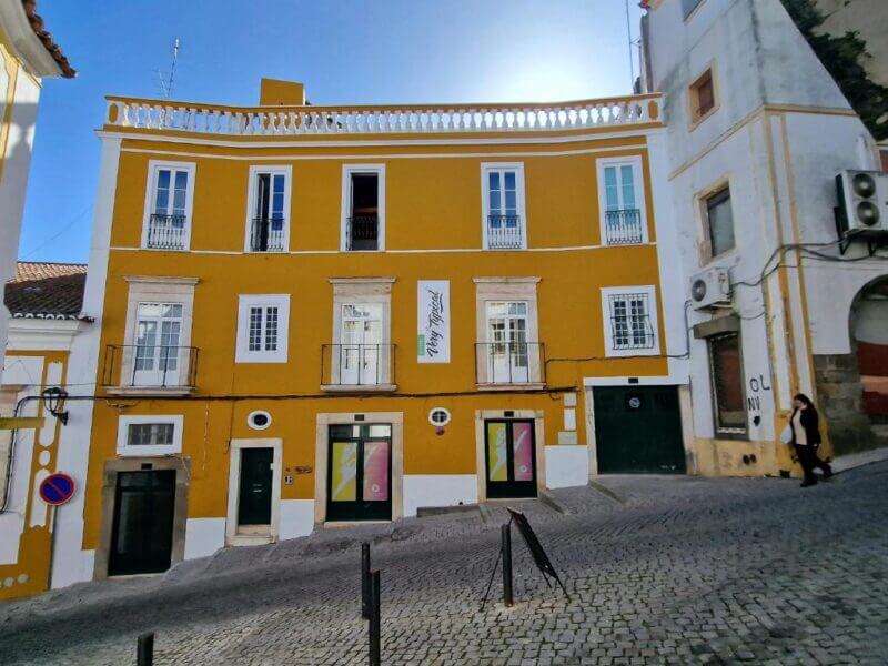 A yellow three-story building with white trim stands on a cobblestone street with a steep incline in Elvas; sunlight shines from behind the roof as a woman walks by this charming Alojamento Local.