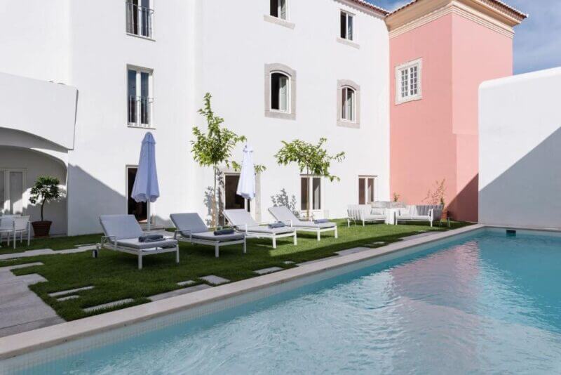 Outdoor pool area with white lounge chairs and umbrellas on a grassy section beside the white and pink Travassos 11 building in Elvas, all set under a clear sky.