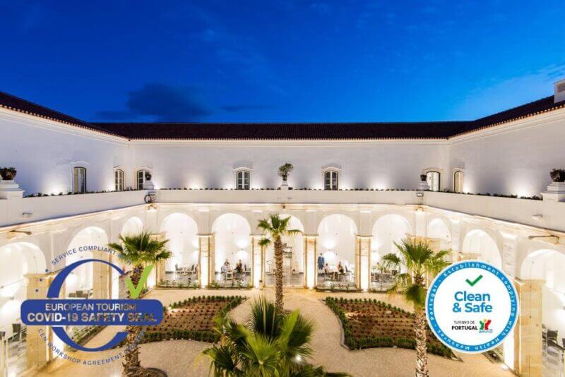 A white, two-story luxury hotel in Portugal surrounds a courtyard with palm trees and outdoor seating at dusk. Two certification seals for COVID-19 safety and cleanliness are displayed at Vila Gale Collection Elvas, a renowned Elvas hotel.