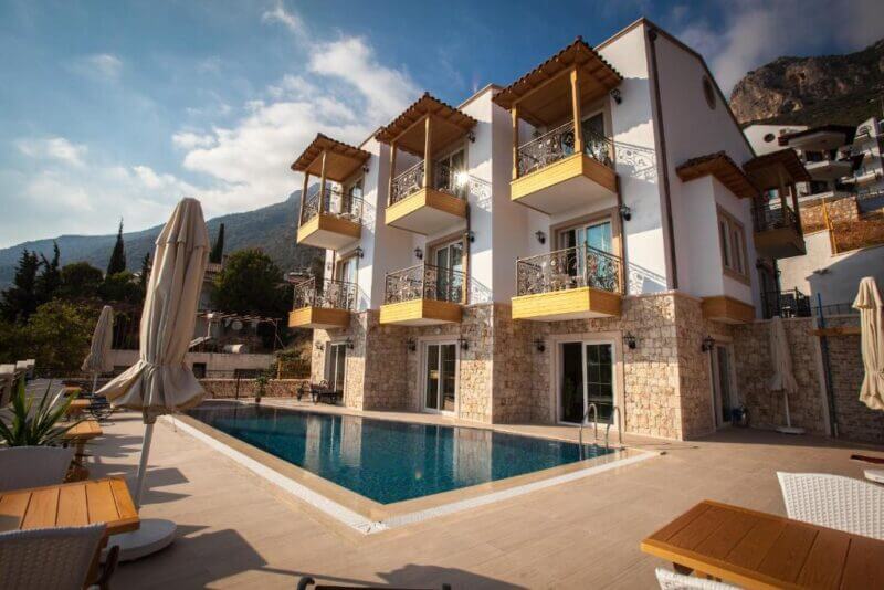 Hotel Aysima in Kas is a modern three-story stone and white building with balconies overlooking a rectangular outdoor swimming pool, surrounded by patio furniture and scenic mountains in the background.