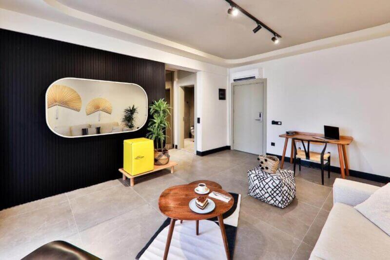 Modern living room at Luff Suites, Kas, with beige tile floor, black accent wall, large oval mirror, yellow mini fridge, small wooden coffee table, pouf, and a wooden desk with a laptop.