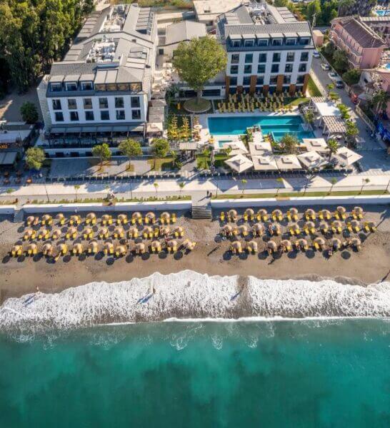 Aerial view of Sundia Exclusive Fethiye, a beachfront hotel with a pool, sun loungers, and umbrellas arranged on the sand facing the ocean.