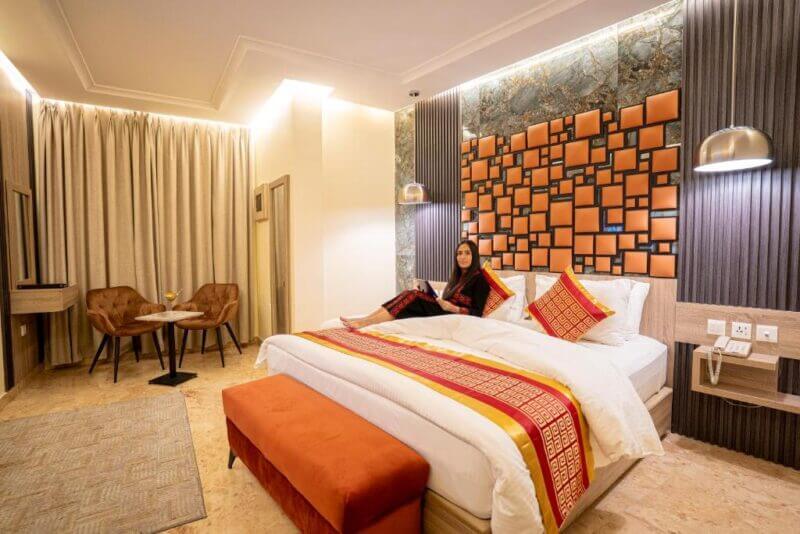 A woman sits on a neatly made bed in a modern room at P Quattro Hotel Petra, with warm lighting, orange accents, and a small seating area with two chairs and a table.