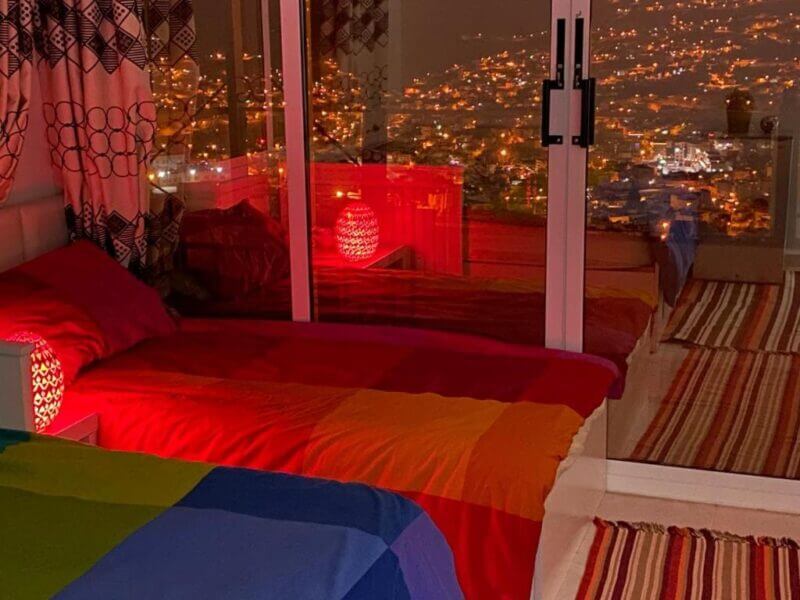 Two single beds with colorful bedding are next to a large glass window at Jordan Guest House, showing Petra’s cityscape sparkling with lights at night; red lamps sit on bedside tables.