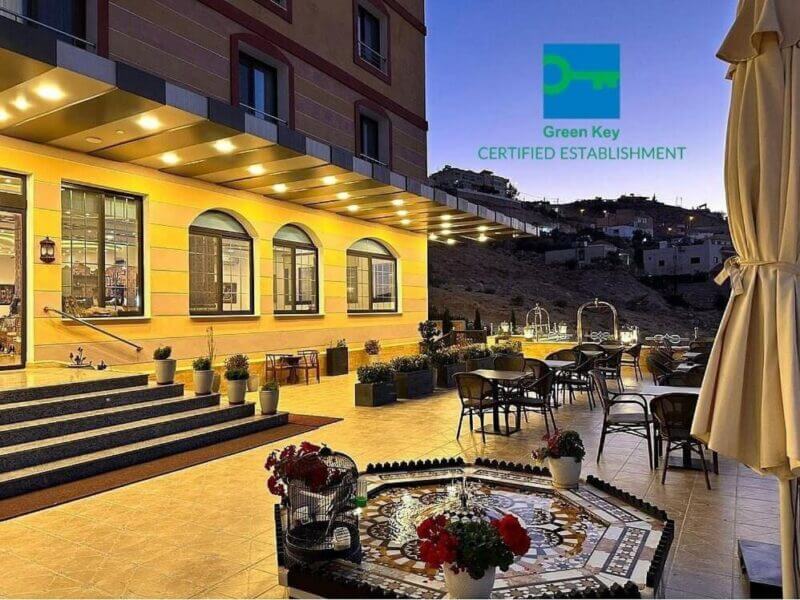 Outdoor hotel terrace at dusk with tables and chairs, a decorative tiled fountain in the foreground, and a Green Key Certified Establishment sign visible—welcome to Canyon Hotel Petra.