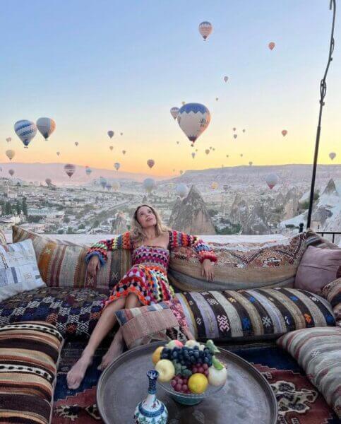 A woman in a colorful dress sits on a patterned outdoor sofa at Arif Cave Hotel in Goreme, with hot air balloons floating over the rocky landscape at sunrise; a table with fruit is in front of her.