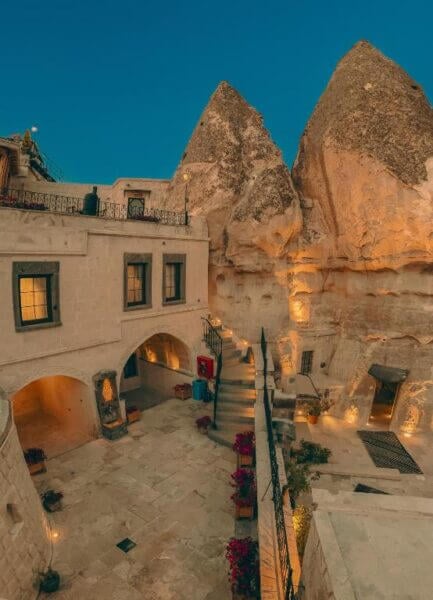 A stone building with arched windows and stairs sits in the Sora Cave area of Goreme, framed by illuminated rock formations at dusk. Potted flowers decorate the courtyard.