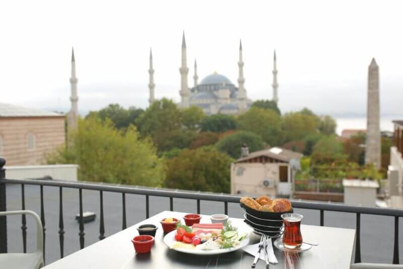 A rooftop table at HaciBayramHotel is set with a Turkish breakfast—tea, cheese, vegetables, and bread—overlooking the Blue Mosque and Istanbul cityscape.