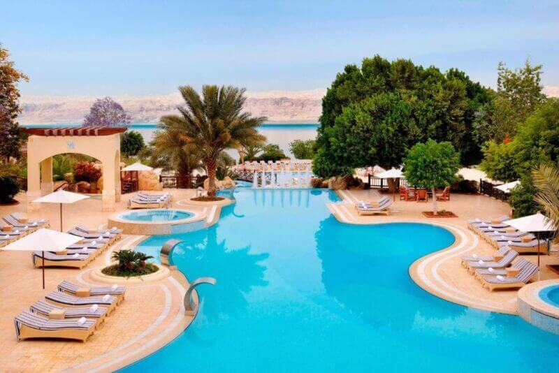Large outdoor swimming pool at this Marriott Resort Dead Sea hotel, surrounded by lounge chairs, umbrellas, and trees, with stunning views of distant mountains and the shimmering Dead Sea under a clear sky.