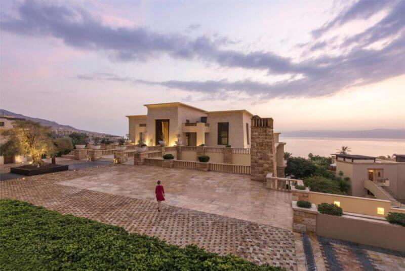 A person in a red outfit walks on a stone courtyard in front of a modern villa at the Kempinski Hotel, overlooking the Dead Sea at sunset.