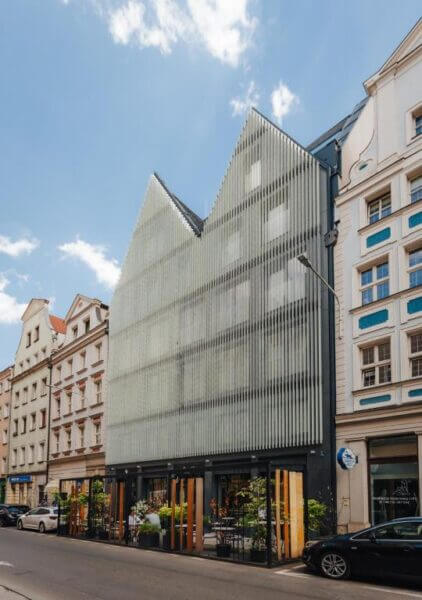 Odrzańska Residence in Wroclaw is a modern building with a glass facade featuring vertical slats and peaked rooflines, standing between traditional buildings on a city street.