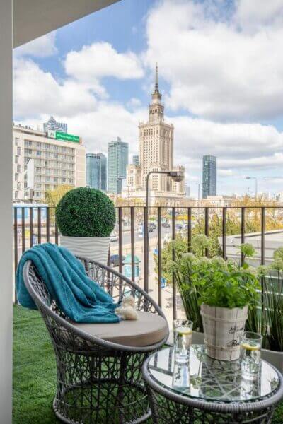 Wicker chair and table with plants on a balcony at Glam Apartments, overlooking Warsaw’s cityscape with a tall, historic building and modern skyscrapers under a partly cloudy sky.