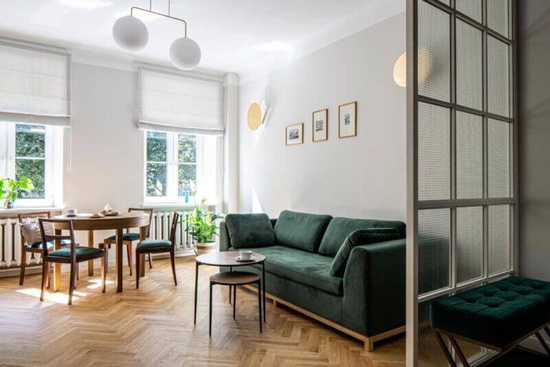 Bright living room in a Heritage Apartment in Warsaw featuring a green velvet sofa, round wooden dining table with chairs, large windows, plants, and minimalist decor.
