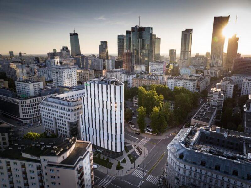 Aerial view of a modern cityscape with high-rise buildings, including the Holiday Inn hotel, tree-lined streets, and a mix of older and contemporary architecture in Warsaw at sunset.