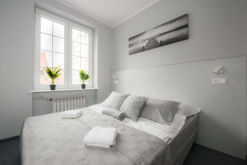 A neatly made double bed with gray and white bedding in a small, light-filled bedroom with two windows, two potted plants, and a black-and-white Wide Street Gdansk photo above the bed.
