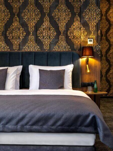 A neatly made bed with white linens and gray blanket, two pillows, patterned gold and black wallpaper, a wall lamp, and a side table with a water bottle create a cozy ambiance at Old Market Residence hotel in Krakow.
