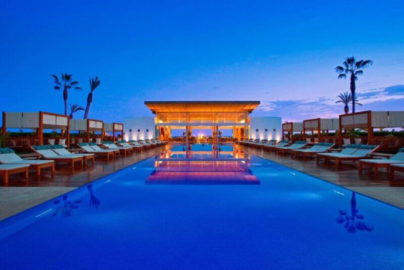 Outdoor swimming pool lined with lounge chairs and cabanas at Hotel Paracas, reflecting a modern pavilion at sunset, with palm trees and a clear blue sky in the background.