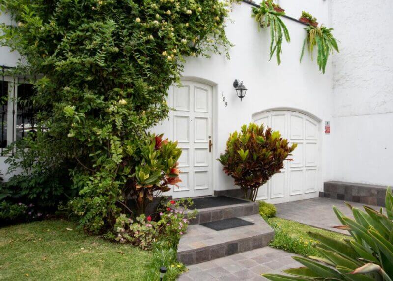 White house entrance with two white doors, green plants and bushes, and hanging ferns above; a paved walkway leads to the door, evoking the inviting charm of a boutique hotel Lima setting.