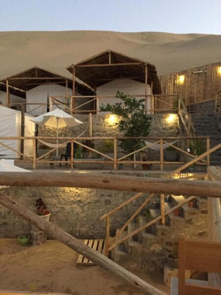 Wooden cabins and round white tents offer desert glamping at EcoCamp Huacachina, set against a sandy hill with fencing, stone walls, stairs, a white umbrella, and outdoor lighting at dusk.