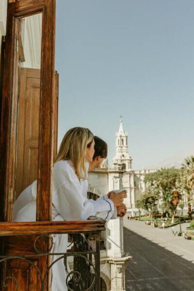 Two people in white robes stand on a balcony, looking out over the city street toward Plaza Central Arequipa, with a church and tower visible in the distance.