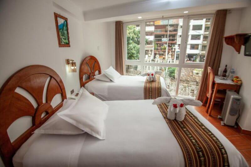 Hotel room with two single beds, decorative bed runners, towel animals, a window with city view reminiscent of Machu Picchu, wall art, and a small table with bottled water and glasses—a cozy Mapiland retreat.