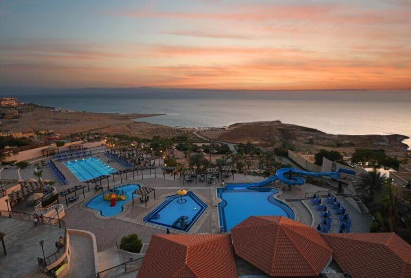 Dead Sea Spa Hotel with multiple swimming pools, water slides, lounge chairs, and a scenic view of the Dead Sea coastline at sunset.