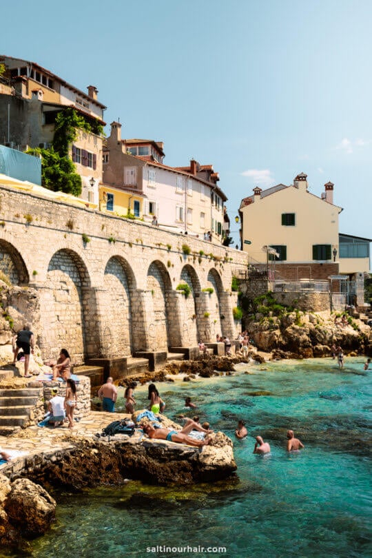 People relax and swim in the clear blue sea at Rovinj Beach, next to stone arches and historic buildings&mdash;a perfect spot on your 3-week itinerary in Croatia.