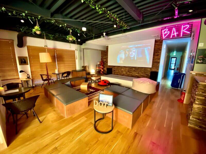 Modern lounge area at Bar Fuji with wooden floors, cushioned seating, coffee tables, a projector screen, and a neon BAR sign. Plants hang from the ceiling and window blinds cover the windows—perfect for relaxing at Hostel Fuji.