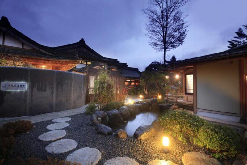 A Japanese-style building at dusk with a stone path, pond, garden plants, and warm exterior lights reminiscent of Onsenji near Fuji.